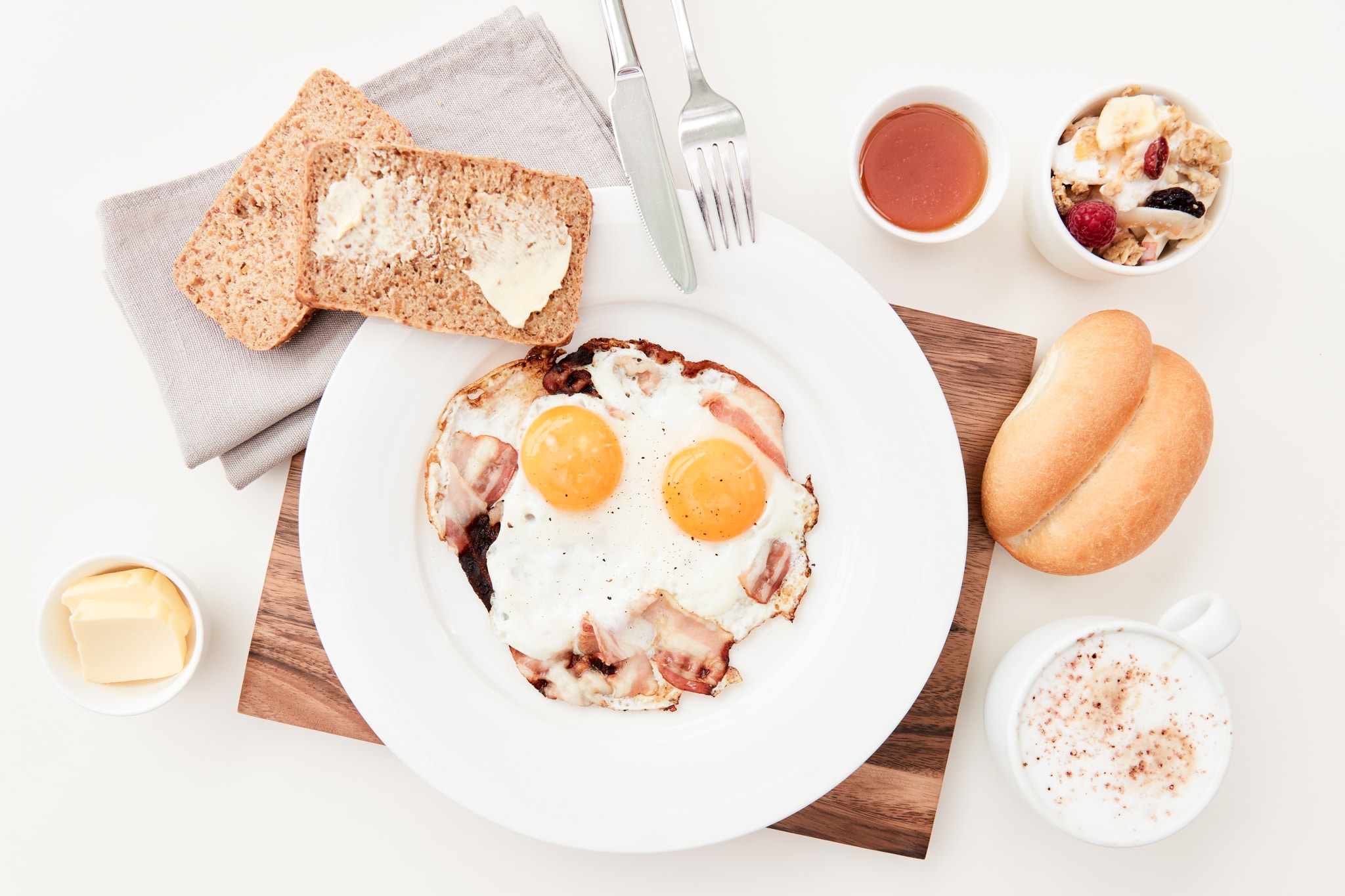 Zu sehen sind Bestandteile eines Frühstücks: ein Teller mit Speck und Spiegelei, daneben Butter, Brot, Kakao, eine Semmel, Marmelade und ein Müsli.