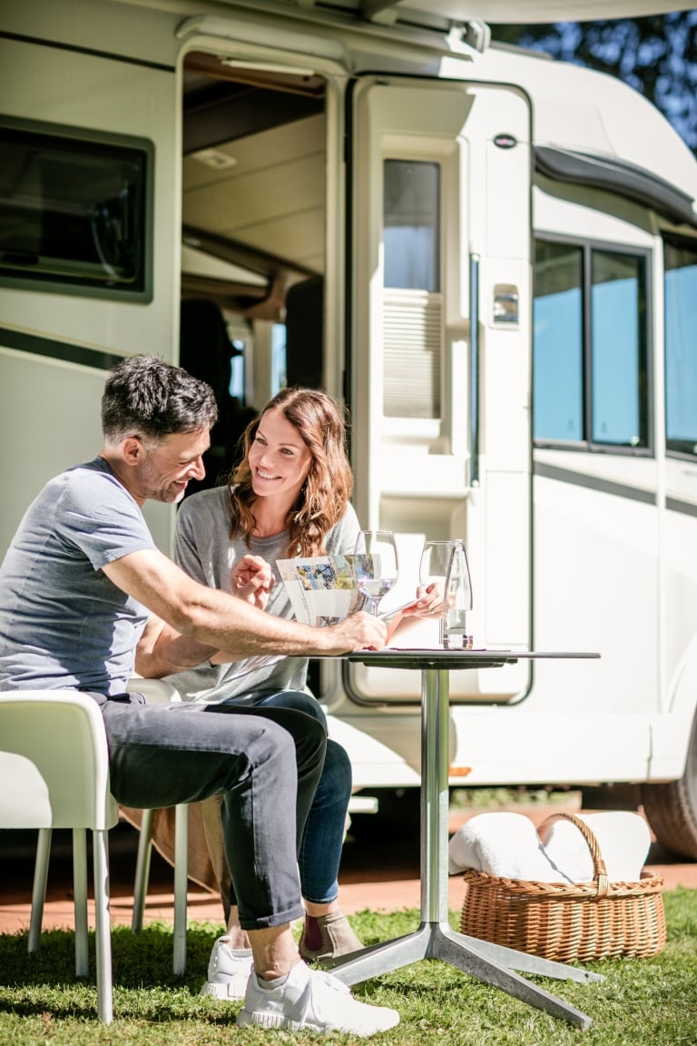 Ein Mann und eine Frau sitzen an einem Camping-Tisch und blättern durch eine Zeitschrift. Im Hintergrund ist ein Wohnwagen zu sehen.