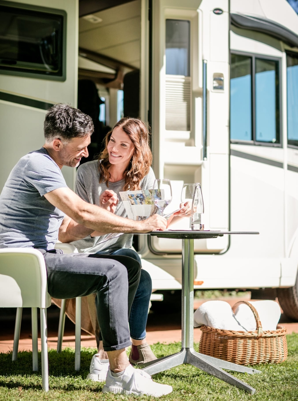 Ein Mann und eine Frau sitzen beim Camping im Sommer an einem Camping-Tisch und blättern durch eine Zeitschrift. Im Hintergrund ist ein Wohnwagen zu sehen.
