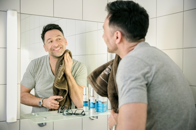 Ein Mann steht im Badezimmer vor dem Spiegel und trocknet sich mit dem Handtuch das Gesicht ab.
