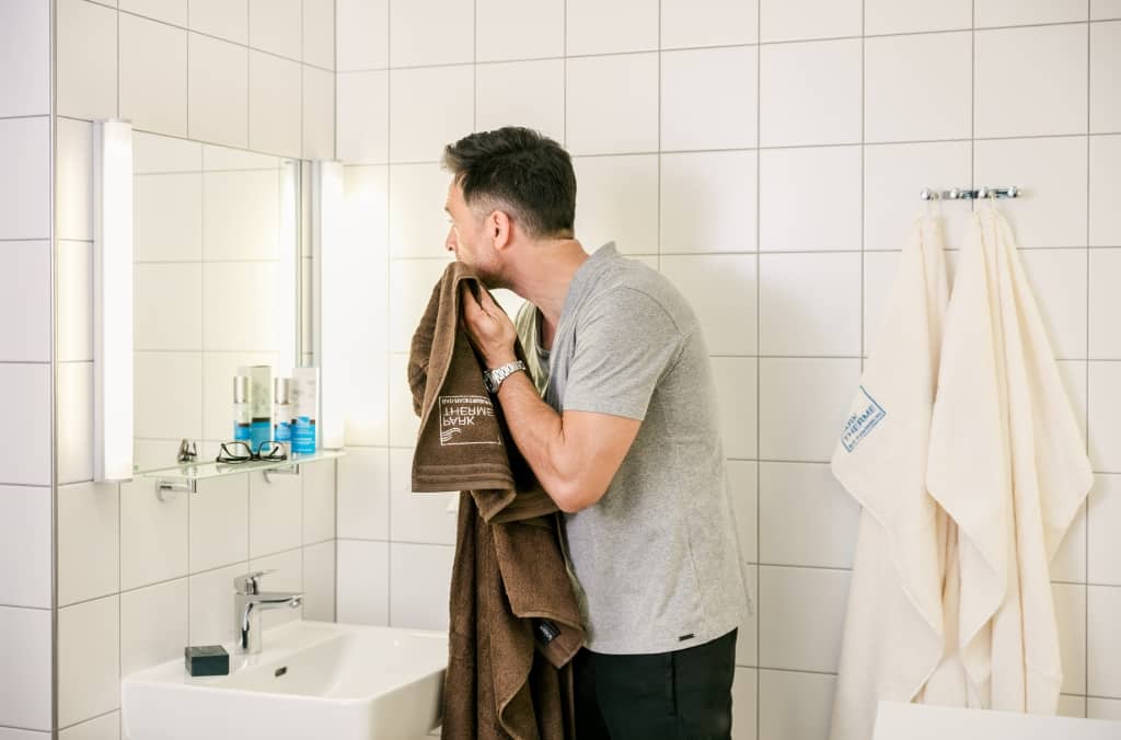 Ein Mann steht im Bad vor dem Spiegel und trocknet sich mit einem Handtuch das Gesicht ab.