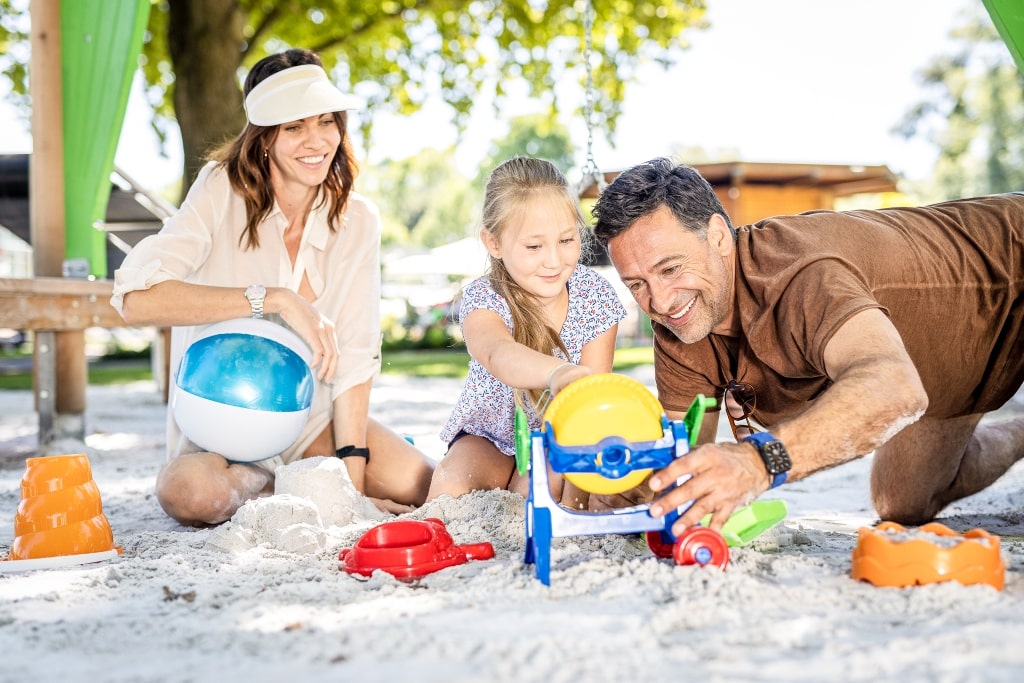 Eine Frau und ein Mann spielen mit einem Kind am Sandspielplatz im Parktherme Fluidum.