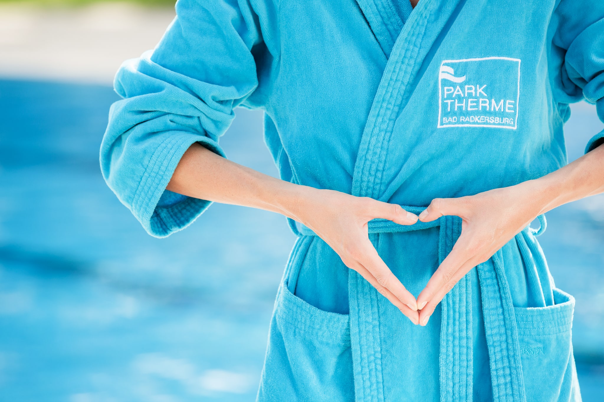 Eine Frau steht im blauen Bademantel vor dem Sportbecken und formt ihre Hände zu einem Herz.