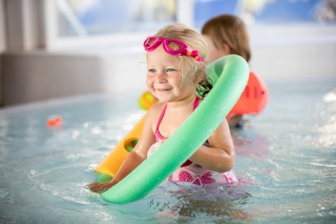 Kleines Kind mit grünem Schwimmreifen um den Hals steht im Babybecken und lächelt.
