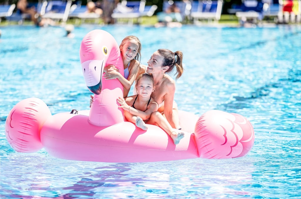 Eine Dame sitzt mit zwei Mädchen auf einem Schwimm-Flamingo im Sportbecken.