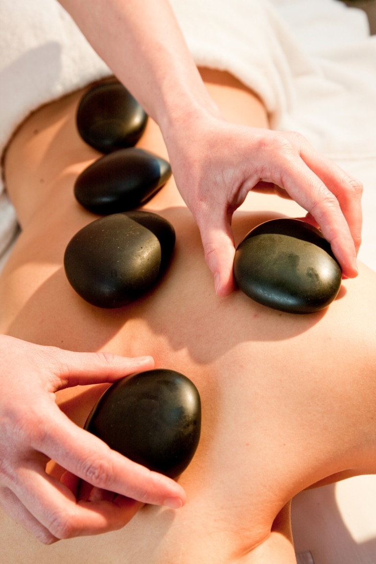 Heiße Steine werden auf dem Rücken einer Dame platziert, die gerade eine Hot Stone Massage genießt.