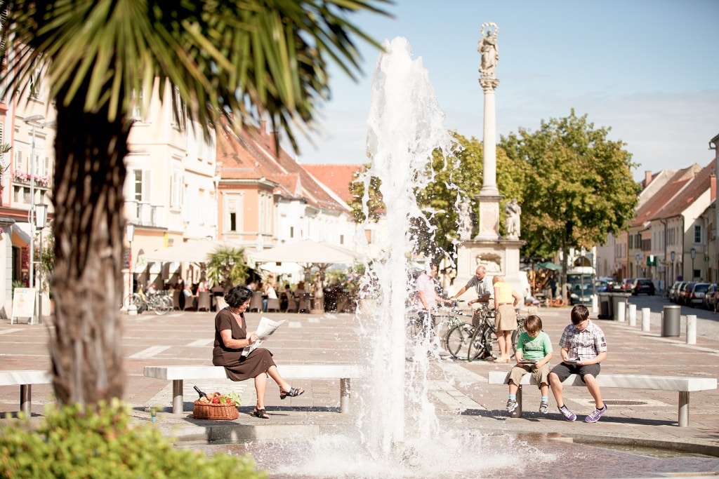 Blick auf den Hauptplatz von Bad Radkersburg. Im Vordergrund ist eine Palme und die Fontäne des Springbrunnens zu sehen, im Hintergrund Personen, die etwas lesen oder in einem Lokal sitzen.