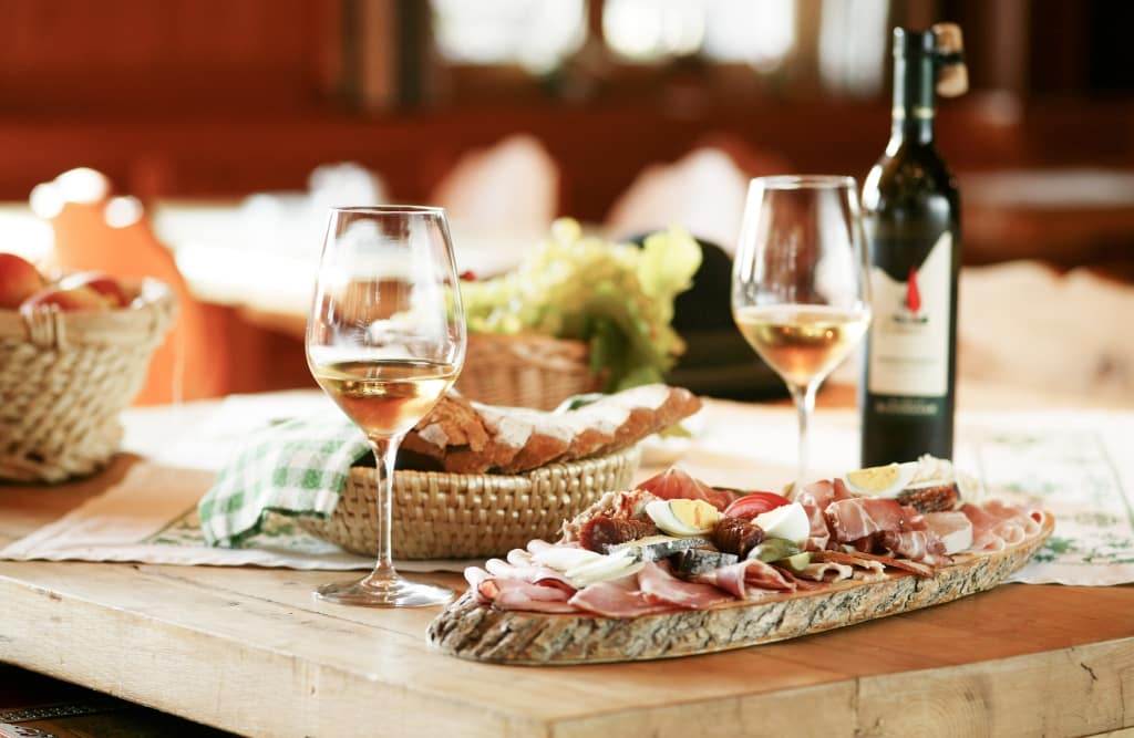 Zu sehen ist ein gedeckter Tisch in einem Buschenschank mit einer Jause. Ein belegtes Brot, zwei Weingläser, eine Weinflasche, ein kleiner Korb mit Brot gefüllt.
