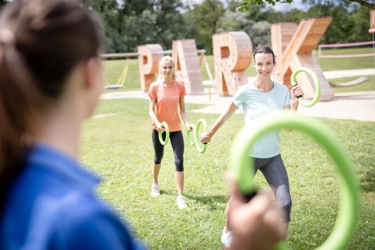 Zwei Frauen stehen mit Smoveys in der Parklandschaft der Parktherme und machen mit einer Physiotherapeutin Übungen.
