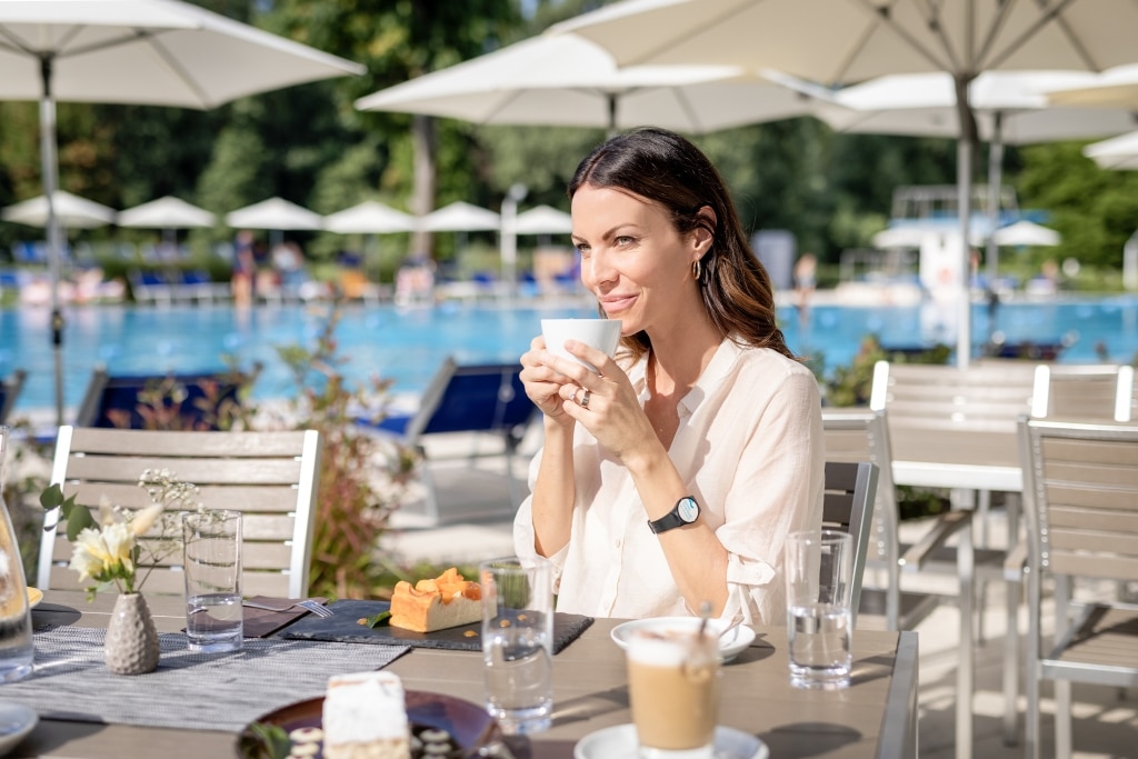 Eine Frau sitzt im Außenbereich des Restaurants Mehrblick und hält eine Tasse Kaffee in der Hintergrund. Auf dem Tisch stehen auch Teller mit Kuchen, im Hintergrund ist das Sportbecken der Parktherme zu sehen.