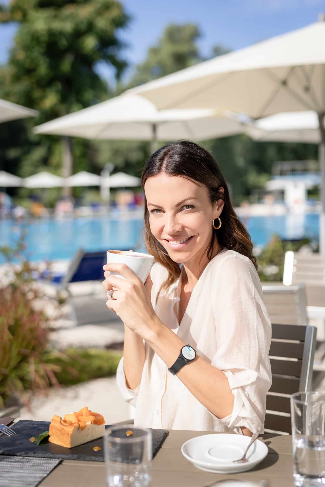 Eine Dame sitzt im Außenbereich des Restaurants Mehrblick und hält eine Tasse Kaffee in der Hand. Im Hintergrund sind Sonnenschirme, Liegen und das 50-Meter-Sportbecken der Parktherme zu sehen.