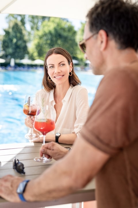 Ein Mann und eine Frau stehen an der Poolbar und stoßen mit einem erfrischenden Getränk an. Im Hintergrund ist das 50-Meter-Sportbecken der Parktherme zu sehen.