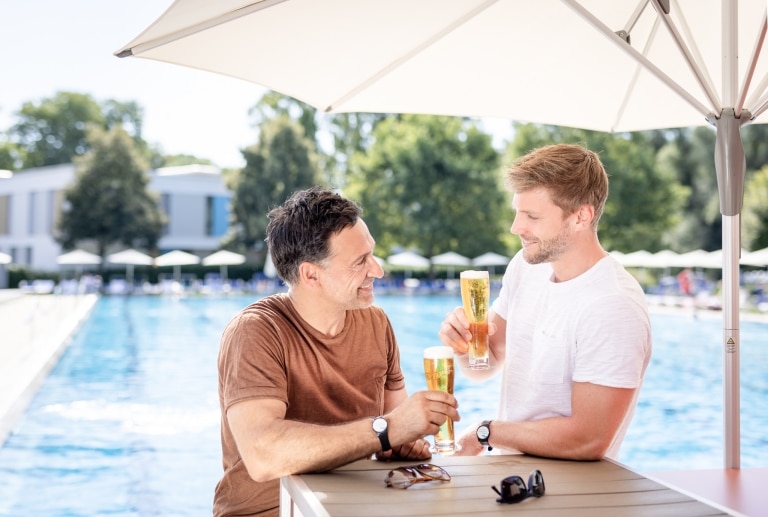 Zwei Männer stehen an einem Tisch an der Poolbar und stoßen mit einem Glas Bier an. Im Hintergrund ist das 50-Meter-Sportbecken der Parktherme zu sehen.