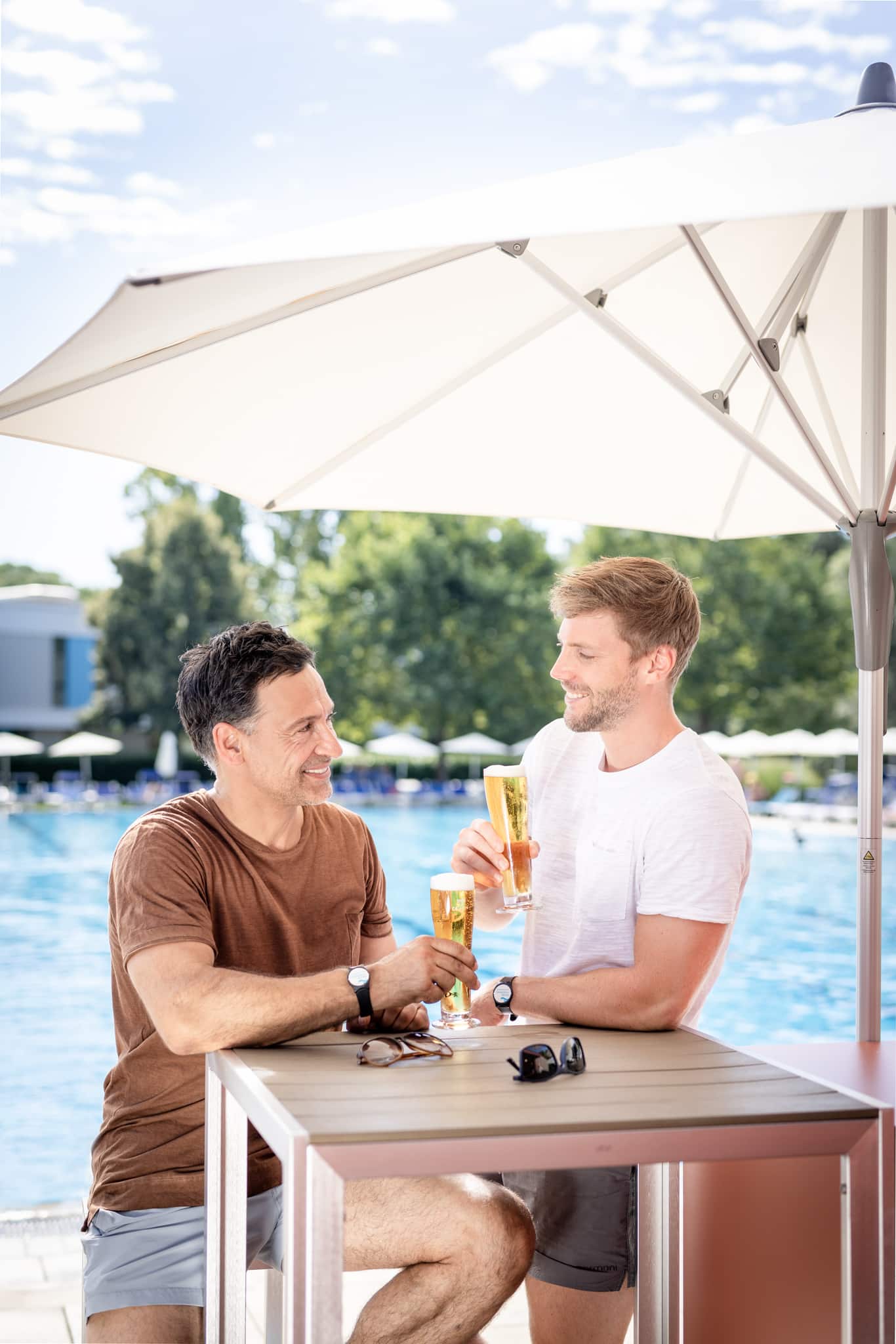 Zwei Männer stehen bzw. sitzen an einem Tisch vor der Poolbar und stoßen mit einem Glas Bier an. Im Hintergrund ist das 50-Meter-Sportbecken der Parktherme zu sehen.