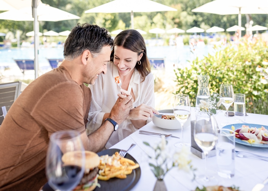 Ein Mann und eine Frau sitzen im Außenbereich des Restaurants Castello und essen. Der Mann hält der Frau eine Gabel mit einem Stück Schinken hin.
