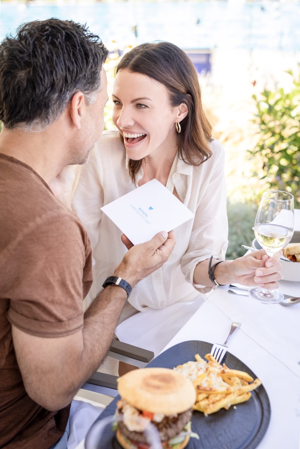 Ein Mann und eine Frau sitzen an einem gedeckten Tisch und essen Burger. Er hält ihr ein Kuvert mit einem Gutschein entgegen.