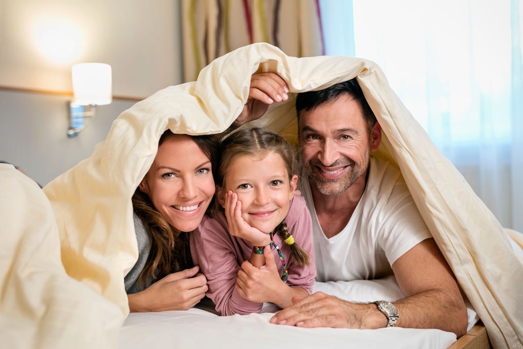 Ein Kind und seine zwei Eltern liegen auf einem Bett und blicken aus der nach oben gehobenen Bettdecke hervor.