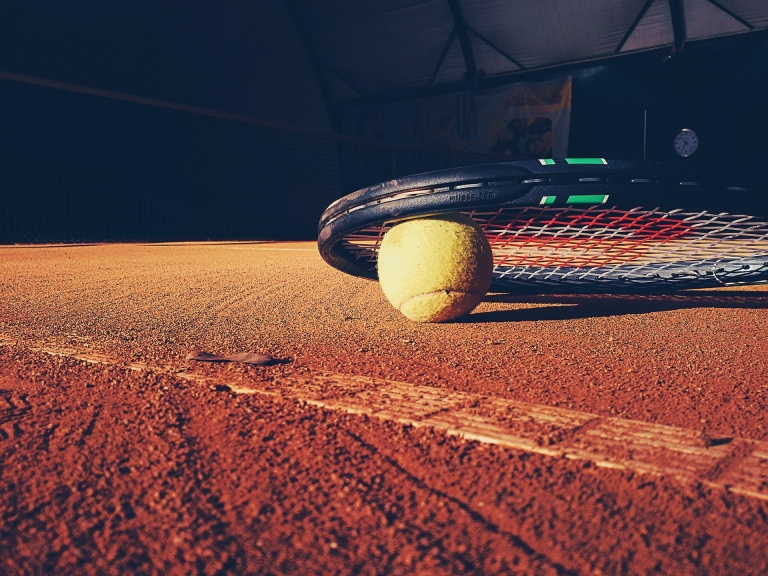 Ein Tennisschläger und ein Tennisball liegen auf einem Sandplatz.