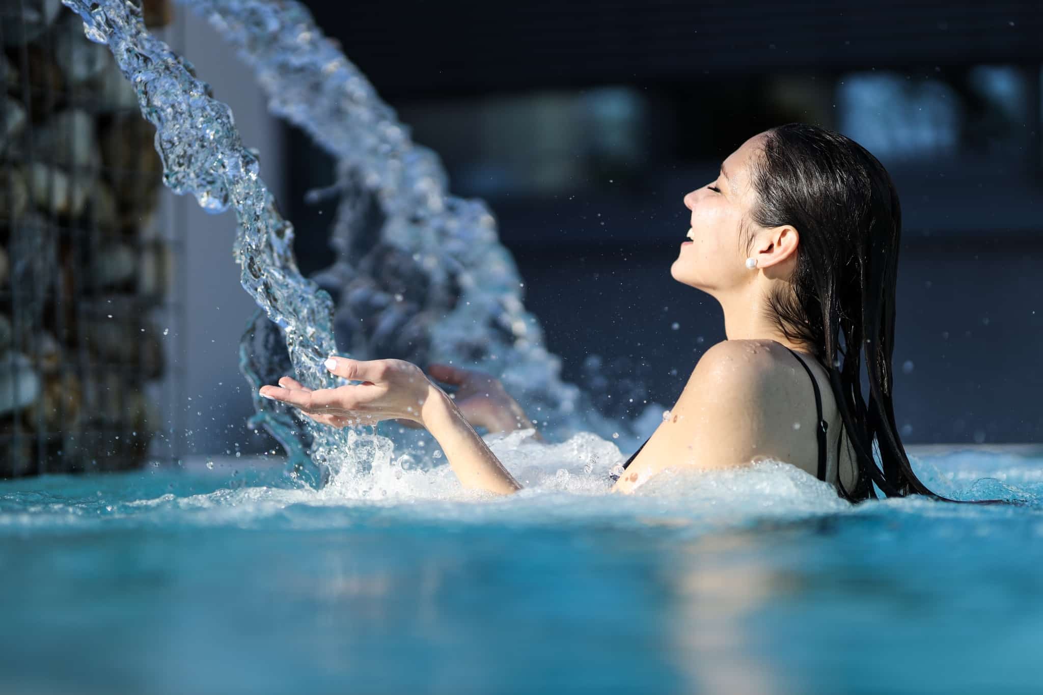 Eine junge Dame steht im 36 Grad heißen Quellbecken der Parktherme und wendet sich dem Wasserfall zu, dessen Wasser ihre beiden ausgestreckten Hände berührt.