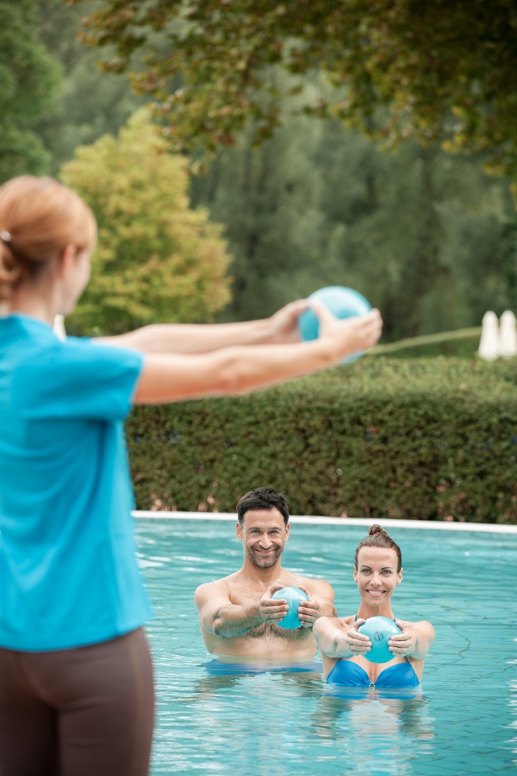 Eine Physiotherapeutin zeigt einem Pärchen im Wasser eine Gymnastikübung mit einem kleinen Ball vor.