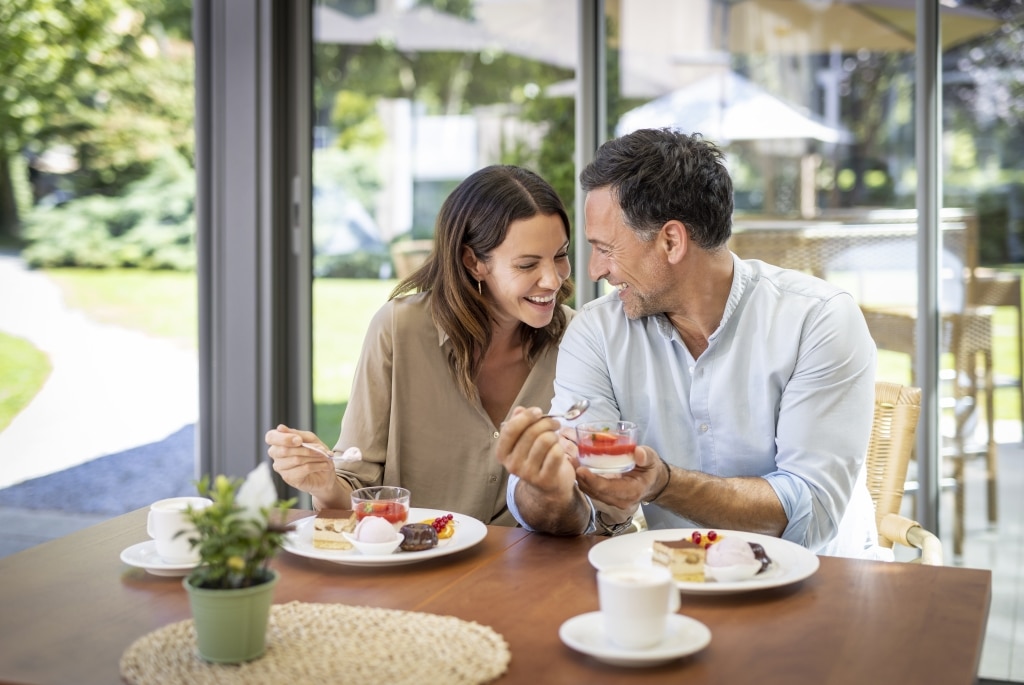 Eine Frau und ein Mann genießen Mehlspeisen und Kaffee im Vitalhotel der Parktherme. Im Hintergrund ist durch das Glas der Garten zu sehen.